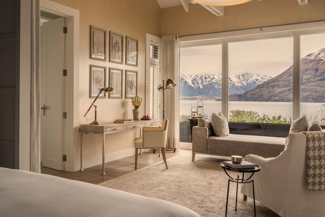 Bedroom suite in a luxury villa with a plush white bed, armchair, desk and day bed, behind the day bed are large windows that look out to the lake and snow-capped mountains.